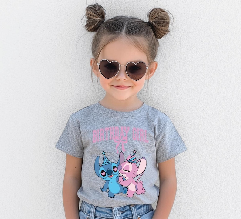 May include: A young girl wearing heart-shaped sunglasses and a gray t-shirt with the text "BIRTHDAY GIRL" in pink. The shirt features a graphic of two cartoon characters, one blue and one pink, wearing party hats.