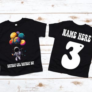 May include: Two black t-shirts, one with a graphic of an astronaut holding balloons and the text "Birthday Girl/Birthday Boy", and the other with the text "NAME HERE" and the number "3" in a retro font.