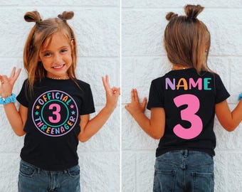 Puede incluir: Una camiseta negra de manga corta con un gráfico circular en la parte delantera con el texto OFFICIAL THREENAGER y el número 3 en el centro. La parte posterior de la camiseta muestra la palabra NAME en letras coloridas sobre un gran número 3 rosa.