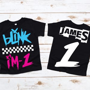 May include: Two black t-shirts with white text and graphics. One shirt has "blink i'm-1" in blue and pink with a checkered pattern. The other shirt has the name "JAMES" and the number "1" in white.