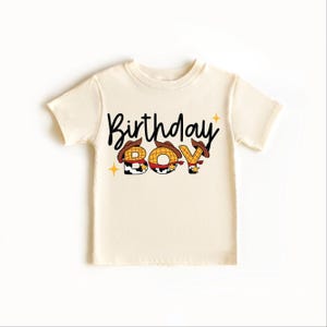 May include: A cream-colored t-shirt with the words "Birthday Boy" in black script. The letters "B", "O", and "Y" are designed with a cowboy theme, featuring a hat and star accents. The shirt is a short-sleeved style.