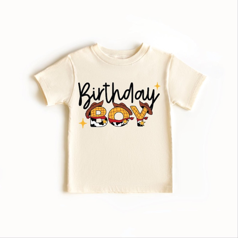 May include: A cream-colored t-shirt with the words "Birthday Boy" in black script. The letters "B", "O", and "Y" are designed with a cowboy theme, featuring a hat and star accents. The shirt is a short-sleeved style.
