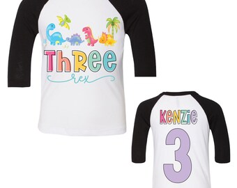 May include: Two white and black baseball shirts with rainbow dinosaur graphics. The first shirt says "Three Rex" and the second shirt says "Kenzie 3".