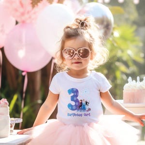 Pode incluir: Uma crian&ccedil;a pequena vestindo uma camiseta branca com o n&uacute;mero 3 azul e um gr&aacute;fico de personagens animados. A camisa tamb&eacute;m diz "Birthday Girl" em azul. A crian&ccedil;a est&aacute; usando um tutu rosa e &oacute;culos de sol cor de rosa. Bal&otilde;es rosa e brancos no fundo.