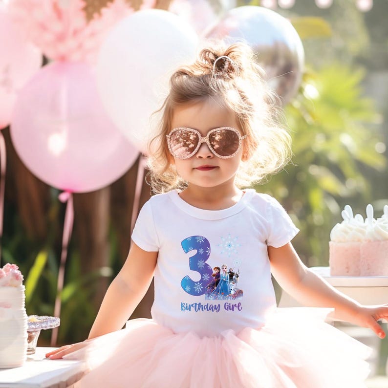 Pode incluir: Uma crian&ccedil;a pequena vestindo uma camiseta branca com o n&uacute;mero 3 azul e um gr&aacute;fico de personagens animados. A camisa tamb&eacute;m diz "Birthday Girl" em azul. A crian&ccedil;a est&aacute; usando um tutu rosa e &oacute;culos de sol cor de rosa. Bal&otilde;es rosa e brancos no fundo.