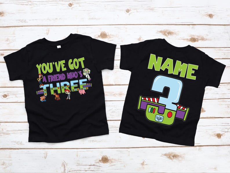 以下が含まれることがあります： カラフルなグラフィックが施された2枚の黒いTシャツ。1枚のシャツには、漫画のキャラクターと一緒に「You've got a friend who's THREE」と書かれています。もう1枚のシャツには、大きな数字の「3」と緑色の「NAME」という文字が書かれています。