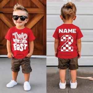 May include: A red short-sleeved t-shirt featuring a white graphic of a race car with the text Two Fast on the front and the word NAME above a large checkered number 2 on the back, paired with brown shorts and white sneakers.