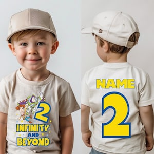 Op de afbeelding: Een beige T-shirt met een cartoon afbeelding van Woody en Buzz Lightyear uit Toy Story. Het shirt heeft het nummer "2" en de tekst "Infinity and Beyond". De achterkant van het shirt heeft het nummer "2" en de tekst "NAME".