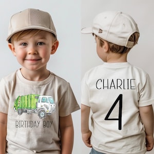 Op de afbeelding: Een beige t-shirt met een aquarel illustratie van een groene en witte vuilniswagen, met de tekst "BIRTHDAY BOY". De achterkant van het shirt heeft de naam "CHARLIE" en het nummer "4" in zwart gedrukt. Het kind draagt een beige baseball cap.