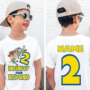 Op de afbeelding: Wit T-shirt met een cartoon afbeelding van Buzz Lightyear en Woody uit Toy Story. Het shirt heeft de tekst "Infinity and Beyond" en het nummer "2" op de voorkant. De achterkant van het shirt heeft de tekst "NAME" en het nummer "2" in geel.