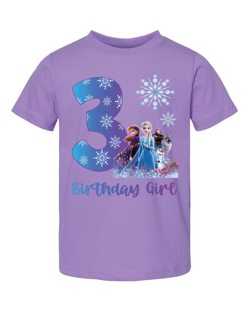 Pode incluir: Uma camiseta lil&aacute;s de manga curta com um gr&aacute;fico do n&uacute;mero 3, flocos de neve e personagens do filme Frozen. O texto "Birthday Girl" est&aacute; abaixo do gr&aacute;fico.