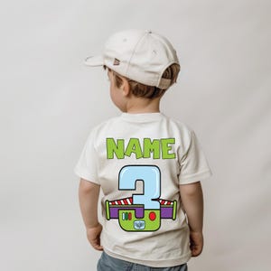 以下が含まれることがあります： クリーム色のTシャツ。緑色のブロック体で「NAME」の文字と、水色、緑、紫、赤で描かれた大きな数字の「3」がプリントされています。ベージュの野球帽をかぶった子供が着ています。