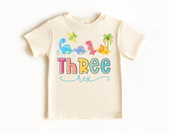 May include: A light beige t-shirt with a rainbow coloured graphic of dinosaurs and the text "Three Rex".