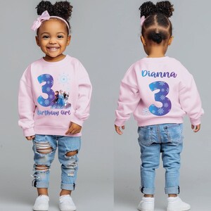 Pode incluir: Um moletom rosa com o n&uacute;mero 3 azul e branco e o texto "Birthday Girl" na frente. O moletom tamb&eacute;m apresenta um gr&aacute;fico de personagens do filme Frozen. A parte de tr&aacute;s do moletom tem o nome "Dianna" e outro 3.