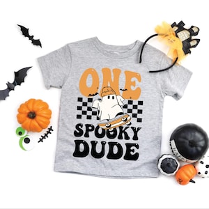 May include: A grey t-shirt with the words "ONE SPOOKY DUDE" in black and orange. The shirt features a cartoon ghost on a skateboard. Halloween decorations, including bats, pumpkins, and a headband, surround the t-shirt.