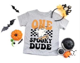 May include: A gray t-shirt with the words "ONE SPOOKY DUDE" in black and orange. The shirt features a cartoon ghost on a skateboard. Halloween-themed decorations, including bats, pumpkins, and a headband, surround the shirt.