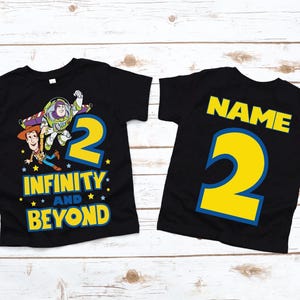 Op de afbeelding: Twee zwarte T-shirts met een Toy Story-thema. E&eacute;n shirt heeft Woody en Buzz Lightyear met het nummer 2 en de tekst "Oneindig en verder". Het andere shirt heeft het nummer 2 en het woord "NAME" in een blauwe omtrek.