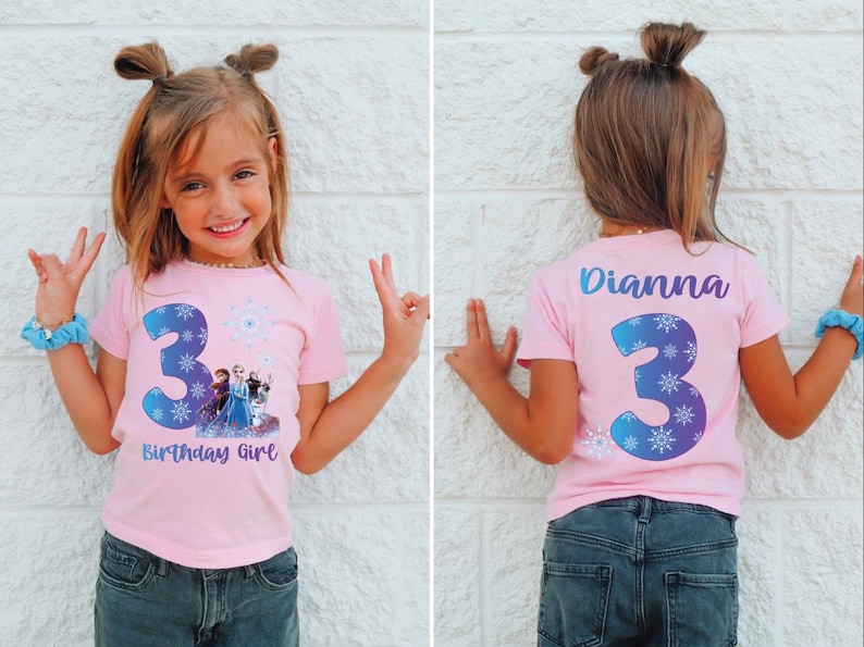 Pode incluir: Uma t-shirt rosa com o n&uacute;mero 3 azul e detalhes de flocos de neve, com personagens de um filme de anima&ccedil;&atilde;o popular. A t-shirt tamb&eacute;m tem o texto "Birthday Girl". A parte de tr&aacute;s da t-shirt tem o nome "Dianna" e o n&uacute;mero 3.