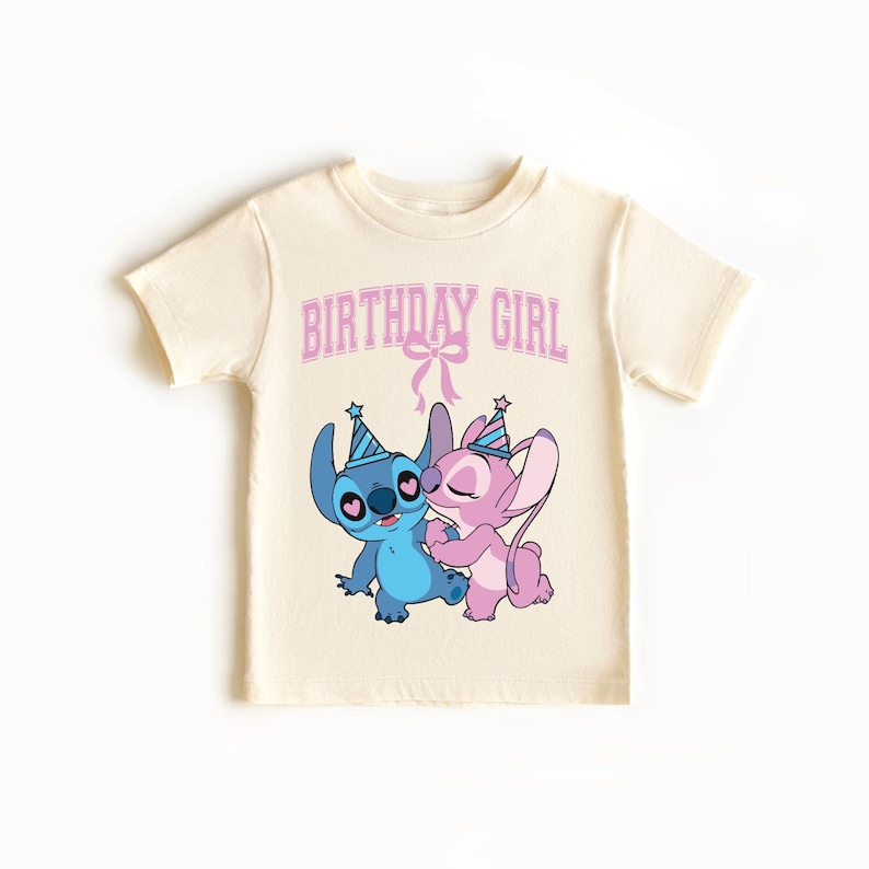 May include: A cream-colored t-shirt featuring a cartoon illustration of two characters, one blue and one pink, wearing party hats and embracing. The text "BIRTHDAY GIRL" is printed above the characters in pink.