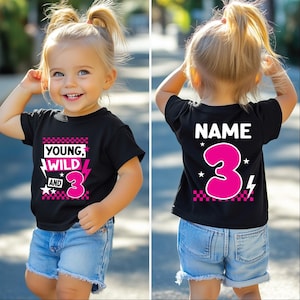 Può includere: T-shirt nera con la scritta "YOUNG, WILD, AND 3" in rosa e bianco, e un numero 3 rosa. La parte posteriore della maglietta ha la scritta "NAME" e un grande numero 3 rosa.