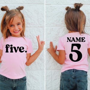 May include: A pink t-shirt with the word "five" in bold black letters on the front. The back of the shirt has the word "NAME" above the number "5". The shirt is worn by a child.