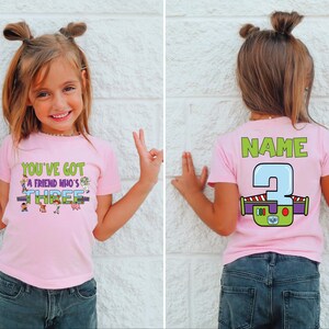 以下が含まれることがあります： 「YOU'VE GOT A FRIEND WHO'S THREE」の文字と漫画のキャラクターがプリントされたピンク色のTシャツ。背中には「NAME」の文字と、バズ・ライトイヤーのデザインの数字「3」がプリントされています。子供は青いジーンズを履いています。