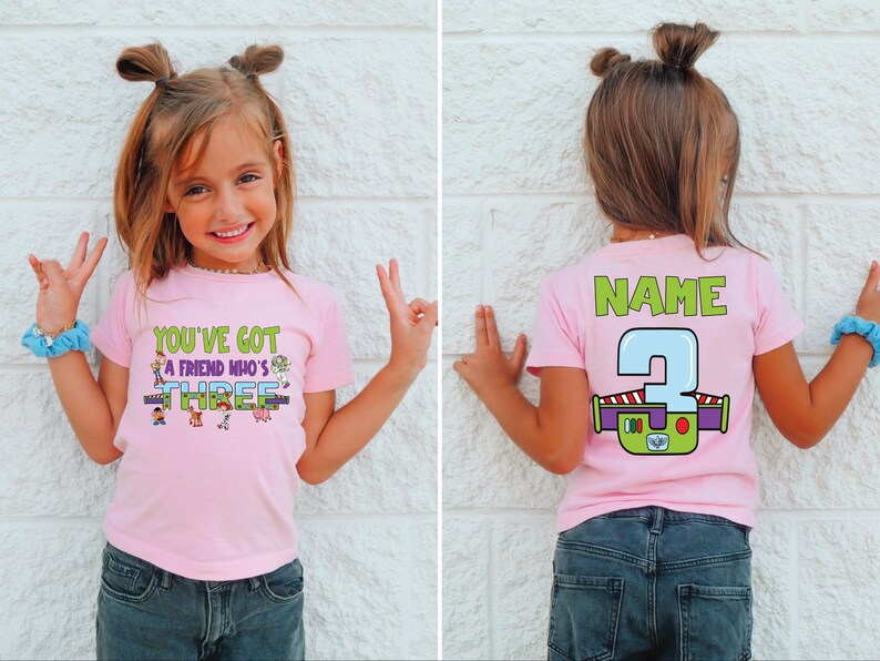 以下が含まれることがあります： 「YOU'VE GOT A FRIEND WHO'S THREE」の文字と漫画のキャラクターがプリントされたピンク色のTシャツ。背中には「NAME」の文字と、バズ・ライトイヤーのデザインの数字「3」がプリントされています。子供は青いジーンズを履いています。