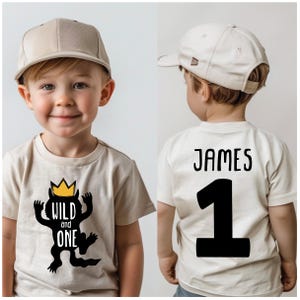 May include: A beige t-shirt with a black and yellow illustration of a monster wearing a crown and the text "Wild and One". The back of the shirt has the name "James" and the number "1" in black.