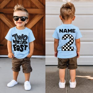 May include: A light blue t-shirt featuring a black graphic with the text Two Fast and a race car on the front, and the text NAME above a large checkered number 2 on the back. The shirt is paired with brown shorts and white sneakers.