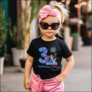 Pode incluir: Uma crian&ccedil;a pequena vestindo uma camiseta preta com o n&uacute;mero 3 azul, flocos de neve e um gr&aacute;fico de personagens do filme Frozen. A camisa tamb&eacute;m tem o texto "Birthday Girl". A crian&ccedil;a est&aacute; usando um la&ccedil;o rosa e &oacute;culos de sol.
