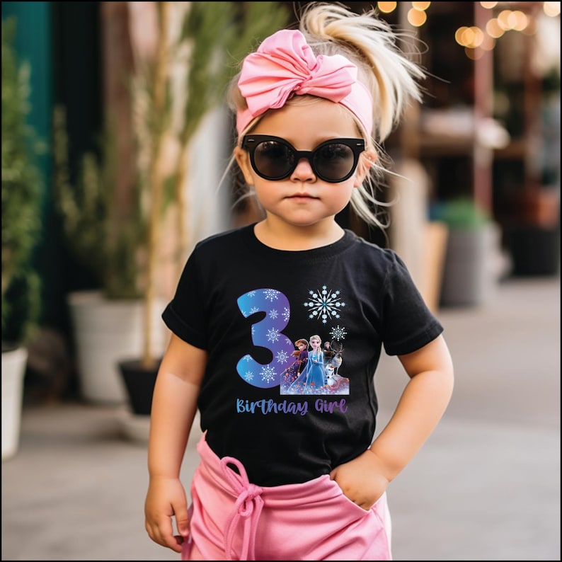 Pode incluir: Uma crian&ccedil;a pequena vestindo uma camiseta preta com o n&uacute;mero 3 azul, flocos de neve e um gr&aacute;fico de personagens do filme Frozen. A camisa tamb&eacute;m tem o texto "Birthday Girl". A crian&ccedil;a est&aacute; usando um la&ccedil;o rosa e &oacute;culos de sol.