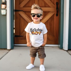 May include: A young child wearing a white t-shirt with the words "Birthday Boy" in a playful font. The shirt is paired with brown shorts and white sneakers. The child is also wearing black sunglasses.