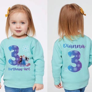 Pode incluir: Camisola azul clara com o n&uacute;mero 3 e a frase "Birthday Girl" na frente. O n&uacute;mero 3 &eacute; decorado com flocos de neve e um design do Frozen. A parte de tr&aacute;s da camisola tem o nome "Dianna" e o n&uacute;mero 3.