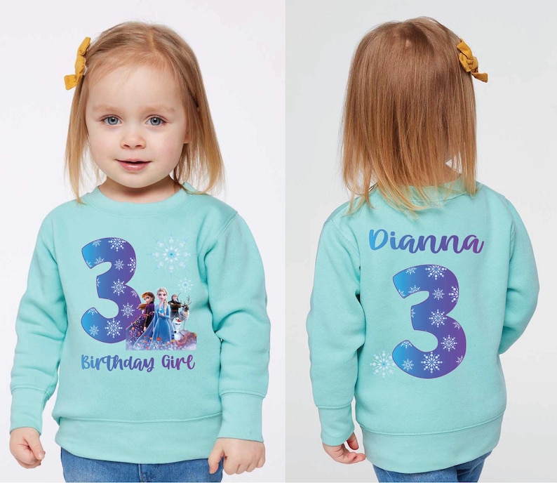 Pode incluir: Camisola azul clara com o n&uacute;mero 3 e a frase "Birthday Girl" na frente. O n&uacute;mero 3 &eacute; decorado com flocos de neve e um design do Frozen. A parte de tr&aacute;s da camisola tem o nome "Dianna" e o n&uacute;mero 3.
