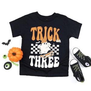 May include: Black t-shirt with the words "TRICK OR THREE" in orange and white. The graphic features a ghost on a skateboard. A small pumpkin, bat, and sneakers are also in the image.