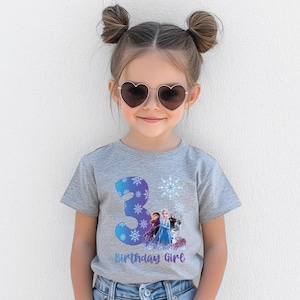 Pode incluir: Uma camiseta cinza com um gr&aacute;fico colorido com o n&uacute;mero 3, flocos de neve e personagens do filme "Frozen". O texto "Birthday Girl" est&aacute; abaixo. A menina est&aacute; usando &oacute;culos de sol em forma de cora&ccedil;&atilde;o.