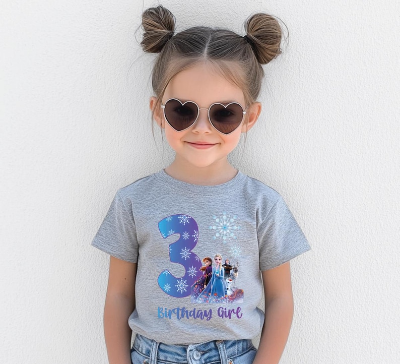 Pode incluir: Uma camiseta cinza com um gr&aacute;fico colorido com o n&uacute;mero 3, flocos de neve e personagens do filme "Frozen". O texto "Birthday Girl" est&aacute; abaixo. A menina est&aacute; usando &oacute;culos de sol em forma de cora&ccedil;&atilde;o.