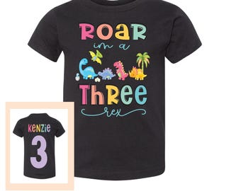 May include: Black t-shirt with a rainbow coloured graphic that says "Roar in a Three Rex". The graphic features a line of dinosaurs. A second black t-shirt with the name "Kenzie" and the number "3" in rainbow colours is shown in the background.