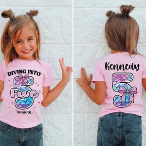 May include: A pink t-shirt featuring the text "Diving Into Five" and a mermaid-scale design for the number 5. The name "Kennedy" is printed below. The back of the shirt also features the name "Kennedy" and a mermaid-scale number 5.