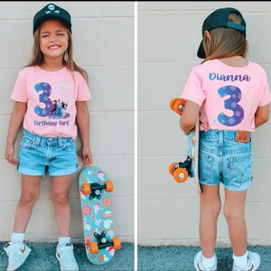 Pode incluir: Camiseta rosa com um grande n&uacute;mero 3 azul e o texto "Birthday Girl". A camisa apresenta uma imagem de personagens do filme Frozen. A menina est&aacute; usando shorts jeans e um bon&eacute; preto, segurando um skate.