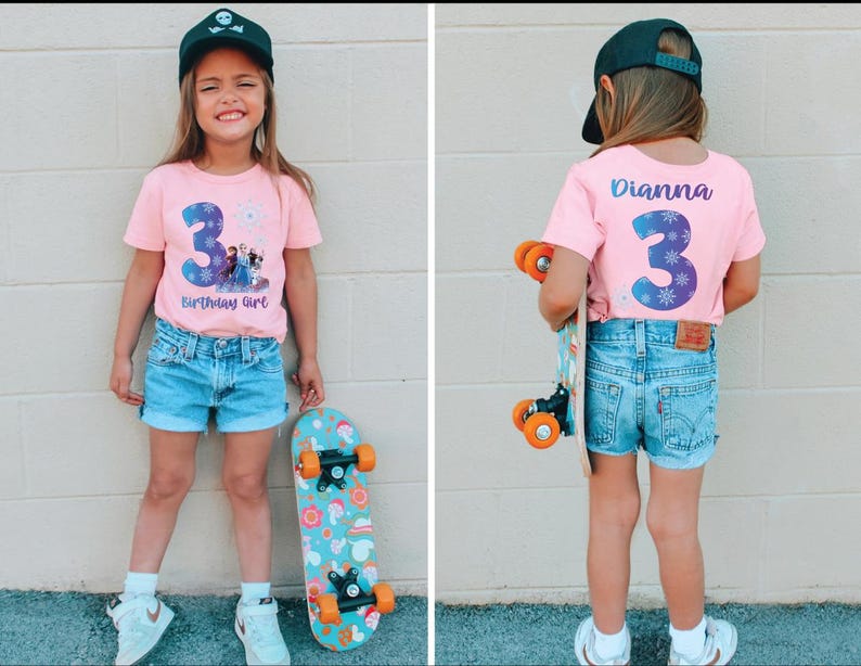 Pode incluir: Camiseta rosa com um grande n&uacute;mero 3 azul e o texto "Birthday Girl". A camisa apresenta uma imagem de personagens do filme Frozen. A menina est&aacute; usando shorts jeans e um bon&eacute; preto, segurando um skate.