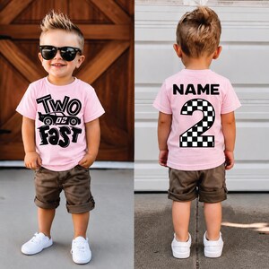 May include: A light pink t-shirt featuring the text TWO FAST with a race car graphic on the front and the word NAME above a large checkered number 2 on the back. The shirt is paired with brown shorts and white sneakers.