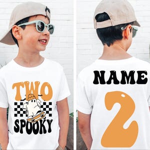 May include: White t-shirt with a Halloween design. The front displays "TWO SPOOKY" in orange and black, with a ghost on a skateboard. The back has the word "NAME" and the number "2" in orange.