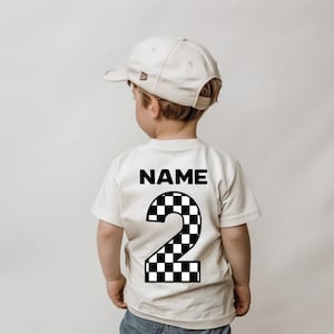 May include: A cream-colored short-sleeve t-shirt featuring the word NAME in black text above a large number 2 with a black and white checkered pattern. The shirt is paired with a plain cream-colored baseball cap worn facing forward.