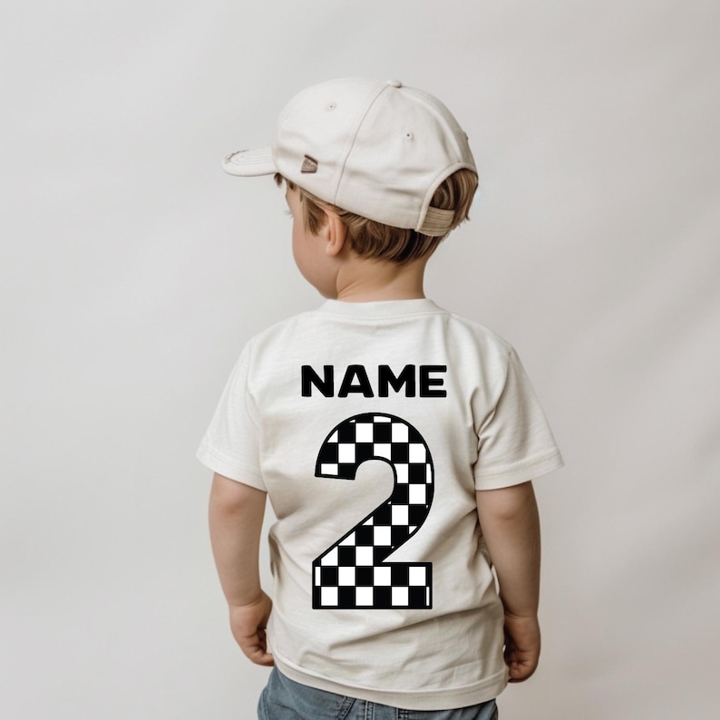 May include: A cream-colored short-sleeve t-shirt featuring the word NAME in black text above a large number 2 with a black and white checkered pattern. The shirt is paired with a plain cream-colored baseball cap worn facing forward.