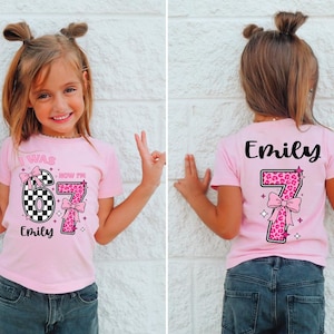 May include: Pink short-sleeved t-shirt with the text "I WAS NOW I'M 7" and the name "Emily". The number 7 is designed with a checkered pattern and a leopard print. The back of the shirt also has the name "Emily" and the number 7.