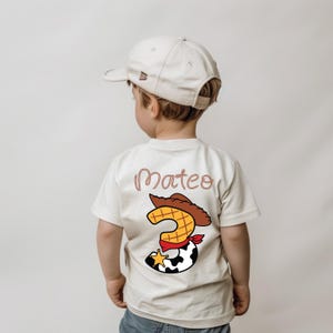 May include: A beige t-shirt with the name "Mateo" in a cursive font. The shirt features a cartoon graphic of a cowboy hat, the number "3", and a cowhide pattern. The child is wearing a beige baseball cap.
