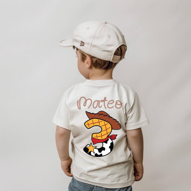 May include: A beige t-shirt with the name "Mateo" in a cursive font. The shirt features a cartoon graphic of a cowboy hat, the number "3", and a cowhide pattern. The child is wearing a beige baseball cap.