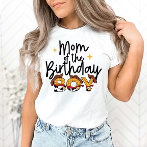 May include: White t-shirt with the text "Mom of the Birthday Boy" in black script. The word "Boy" is designed with cowboy hat graphics. The shirt is a casual style.