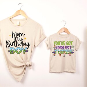 以下が含まれることがあります： 木製のハンガーに掛けられた2枚のベージュ色のTシャツ。1枚のシャツには、黒い筆記体で「Mom of the Birthday Boy」と書かれ、カラフルなアクセントが施されています。もう1枚のシャツには、漫画のキャラクターと一緒に「You've Got a Friend Who's Three」と書かれています。